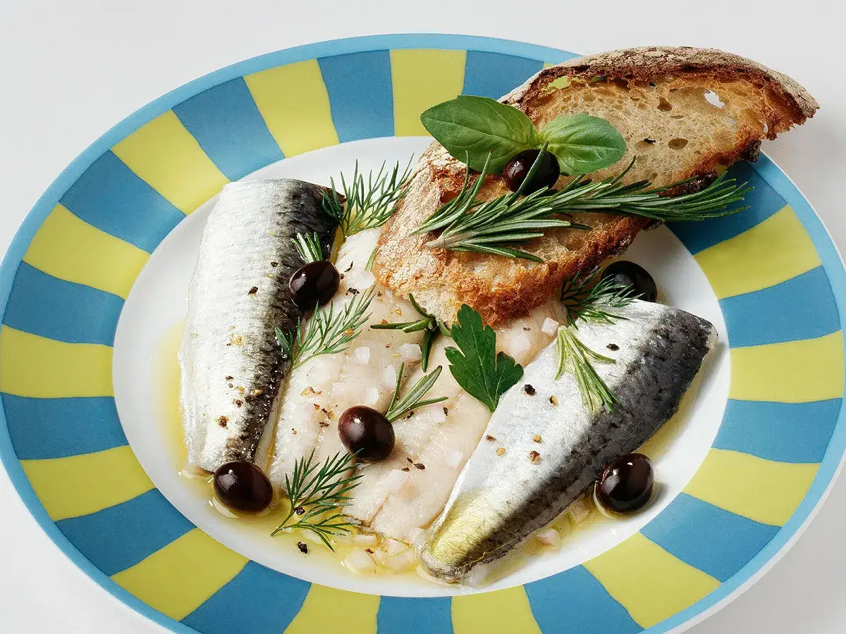 Sardines à l'escabèche, olives noires, herbes fraîches et pain grillé sur une assiette rayée bleu et jaune.