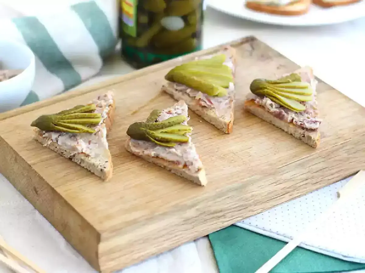 Tranches de pain de campagne garnies de rillettes de porc et de cornichons, pr&eacute;sent&eacute;es sur une planche en bois.